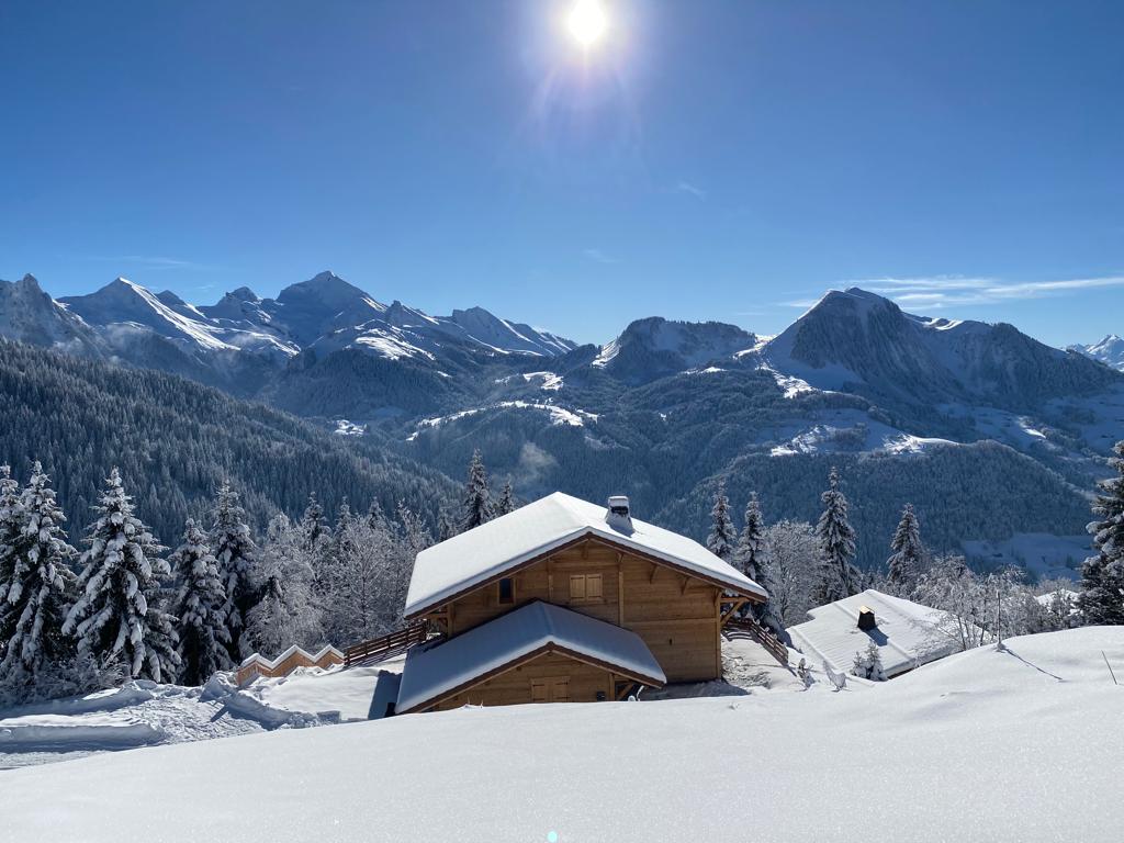 The chalet is set against a backdrop of snow-covered mountains, surrounded by frosted trees. Sunlight glints off the roof, highlighting its wooden structure. A vast expanse of untouched snow blankets the ground, creating a serene winter landscape.