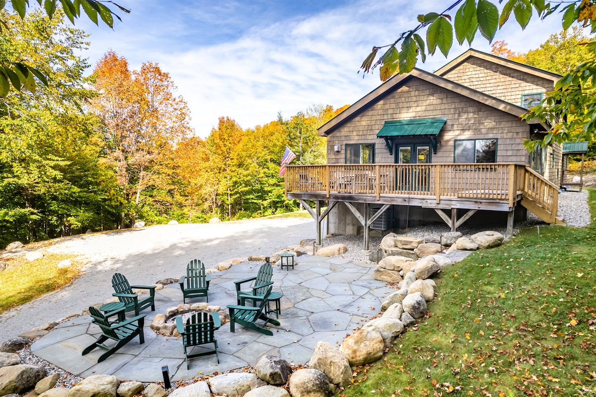 A spacious outdoor patio features a circular arrangement of green Adirondack chairs positioned around a stone fire pit. The house is visible in the background, complemented by a wooden deck and surrounded by vibrant autumn foliage.