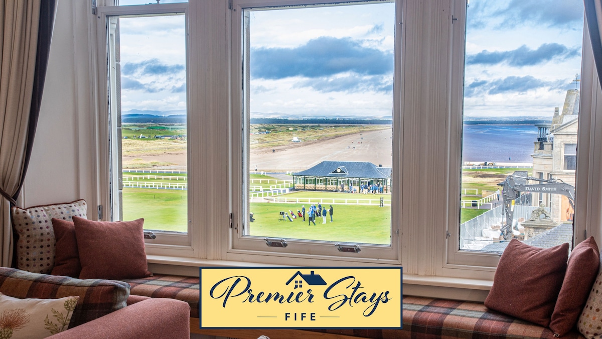 Large windows provide sweeping views of the iconic Old Course and West Sands beach, inviting natural light into the interior. Comfortable cushions on a sofa enhance the space, creating an ideal spot for enjoying the picturesque surroundings.