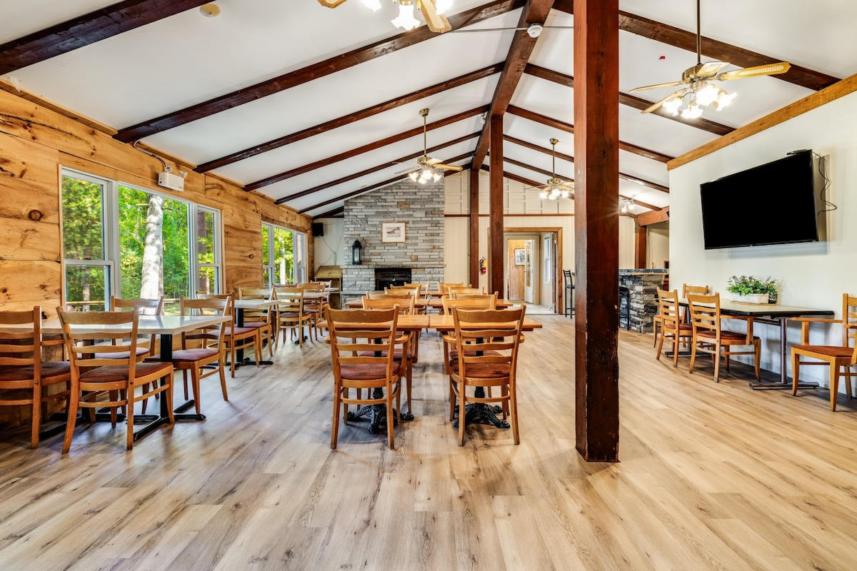 A spacious dining area features numerous wooden tables and chairs arranged to accommodate guests. Wooden beams span the ceiling, and natural light filters through large windows, highlighting the stone fireplace and a flat-screen TV mounted on the wall.