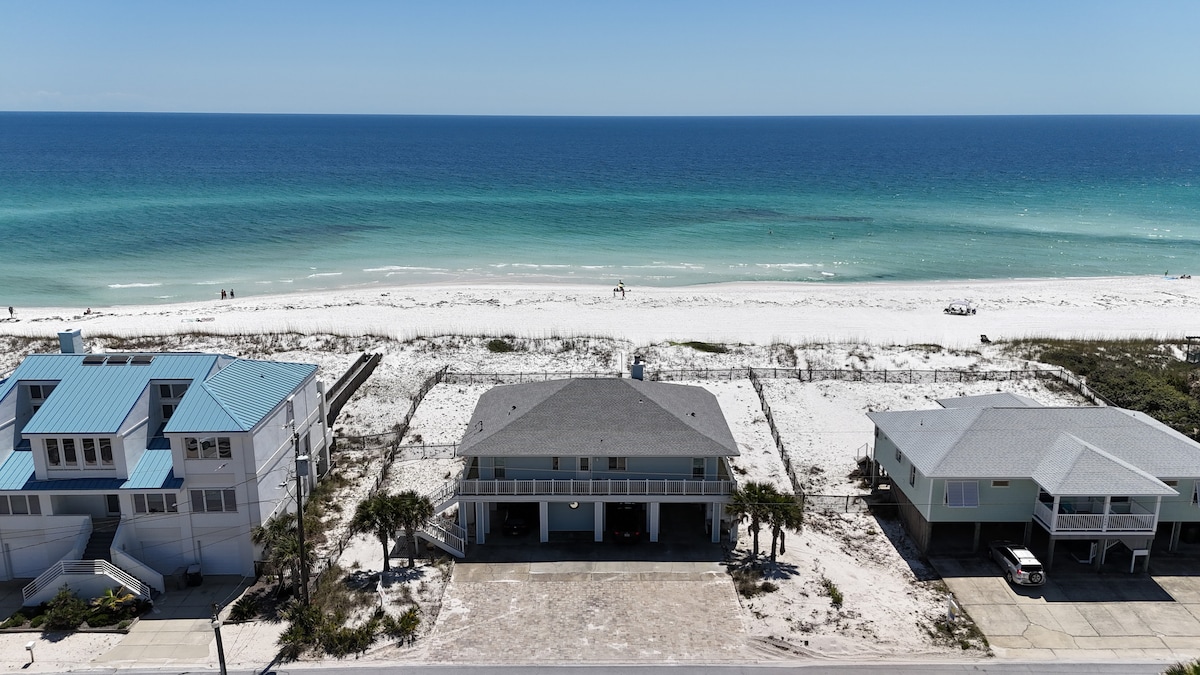 An aerial view captures the coastal landscape, highlighting expansive white sands and the shimmering turquoise Gulf of Mexico. The beachfront property features a spacious yard, framed by palm trees, with neighboring houses positioned on either side along the shoreline.