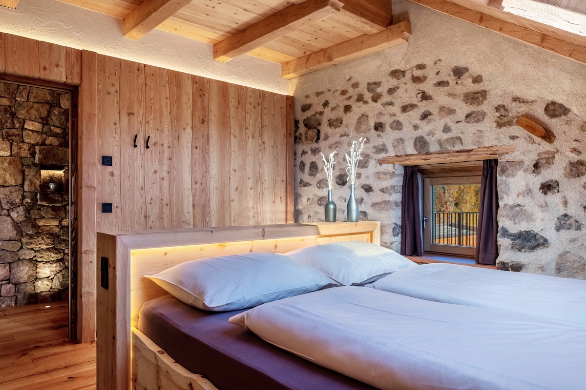 A spacious bedroom features a wooden bed with illuminated accents and two pillows. Natural light enters through a small window, illuminating the stone and wood walls, while a decorative arrangement of vases is positioned on the bedside.