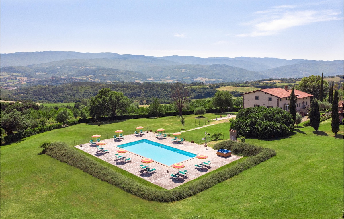 A spacious outdoor area showcases a large swimming pool surrounded by sun loungers and umbrellas. Vibrant green lawns extend beyond the pool, bordered by trees and hills, offering a scenic view of the Tuscan landscape.