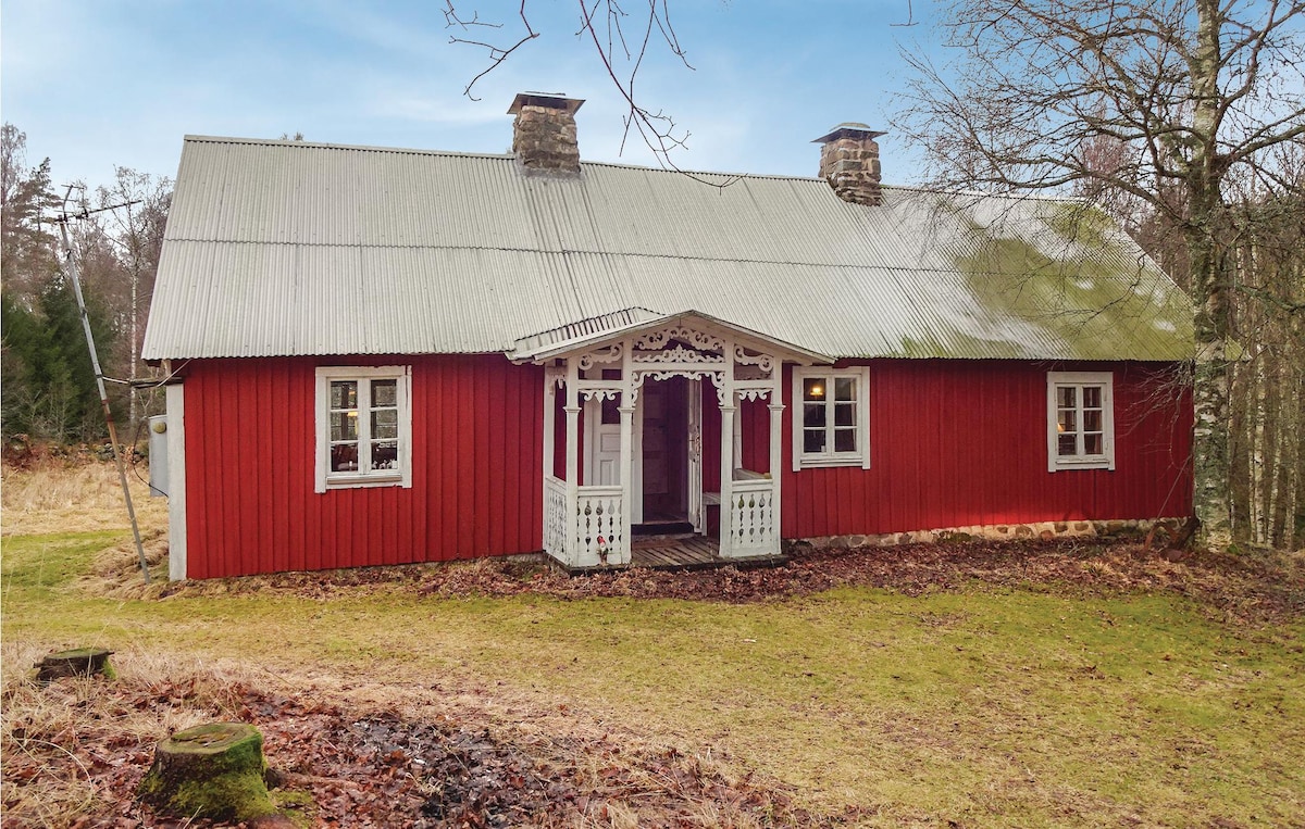 A traditional red wooden house with a metal roof is set against a backdrop of bare trees and grassy land. A decorative porch, featuring intricate woodwork, provides an inviting entrance. The surrounding area showcases a natural, rustic landscape typical of the Swedish countryside.