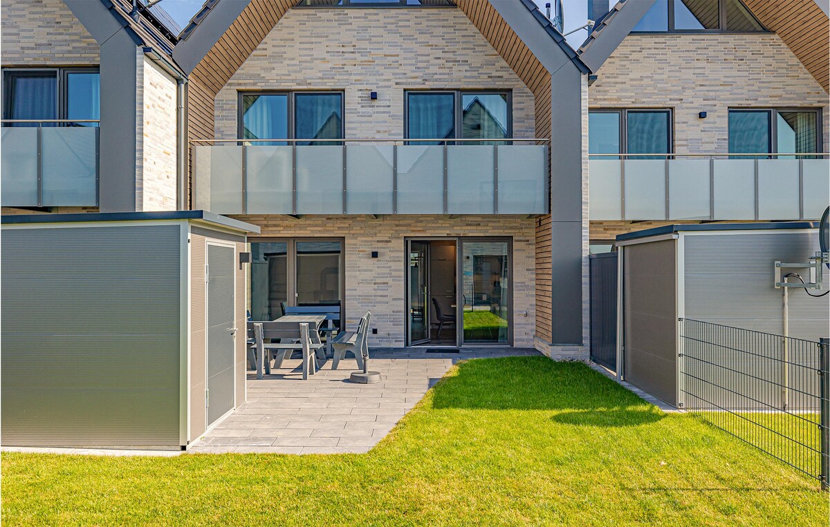 A modern vacation home exterior is presented, featuring a patio area with a rectangular table and four chairs. The building showcases stone and wood accents, with glass railings extending over the upper balconies. A well-maintained lawn adds a touch of greenery to the setting.