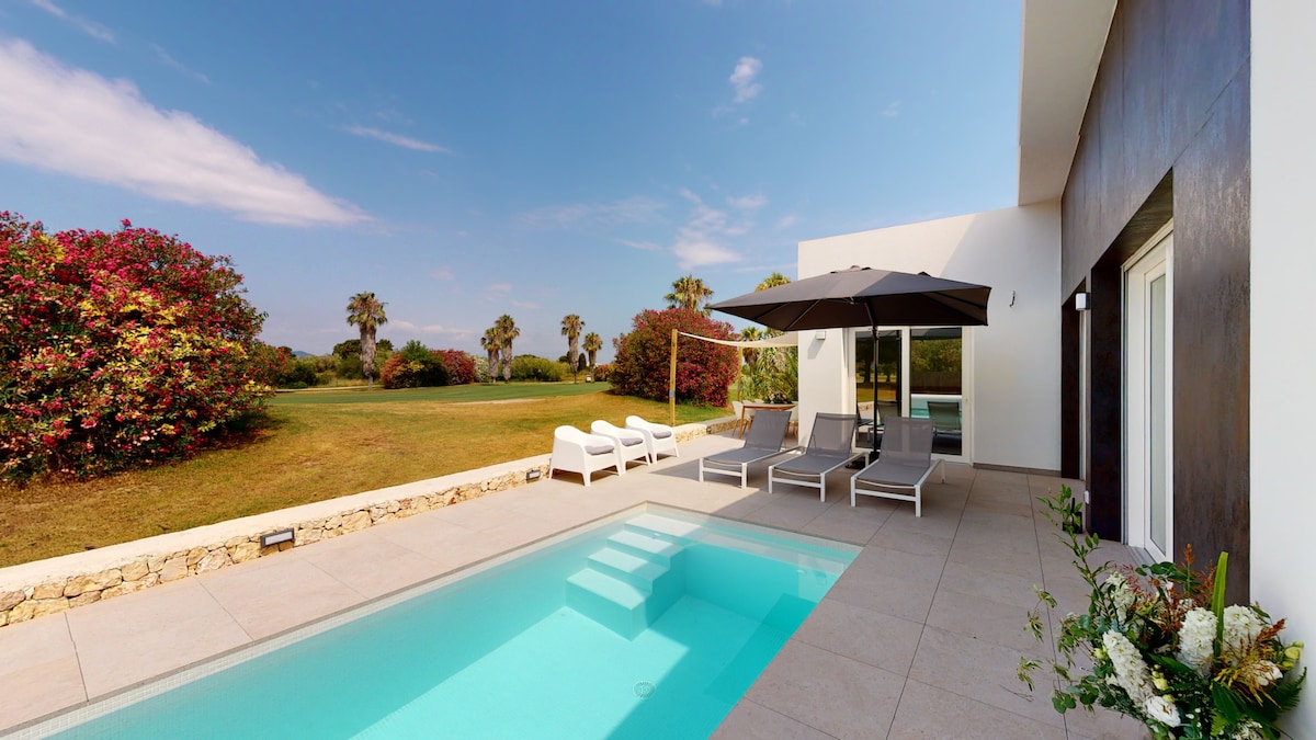 A modern patio area is featured with a swimming pool, surrounded by lounge chairs and an umbrella for shade. Lush greenery and flowering bushes create a scenic backdrop, enhancing the space's relaxing ambiance.
