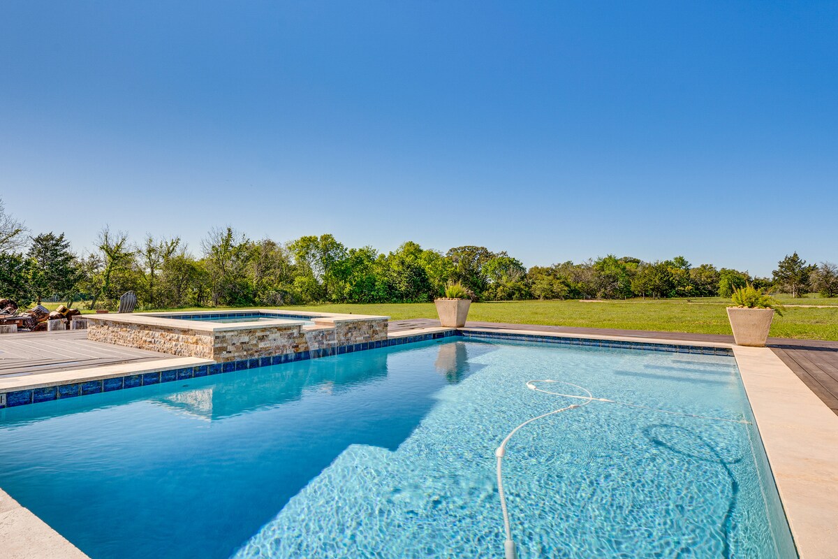 A spacious outdoor pool is surrounded by a wooden deck and overlooks a verdant landscape. The pool features a shallow area and a hot tub, with planters positioned at the pool's edges. A clear blue sky complements the serene setting.