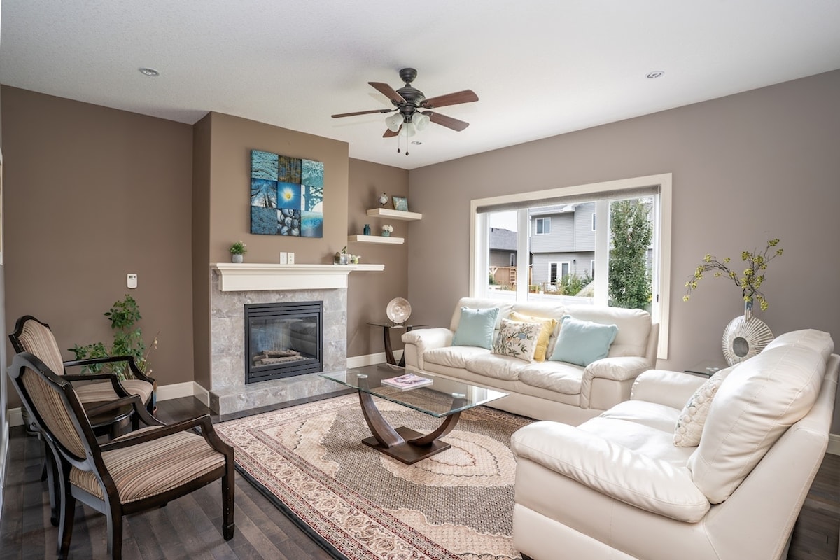 A spacious living room features a light-colored leather sofa and armchairs arranged around a glass coffee table. A beige area rug lies on dark flooring. A fireplace is positioned against a brown accent wall, with artwork displayed above it and a window providing natural light.