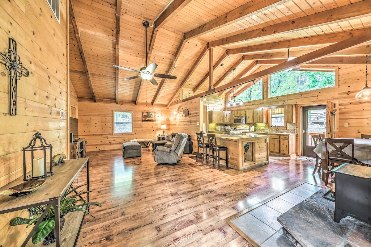 A spacious living area features high wooden ceilings with exposed beams and a ceiling fan. Natural wood paneling surrounds the room. Cozy seating is arranged near a warm-toned stone floor, with a large window overlooking forested views. The kitchen area is open and accessible.