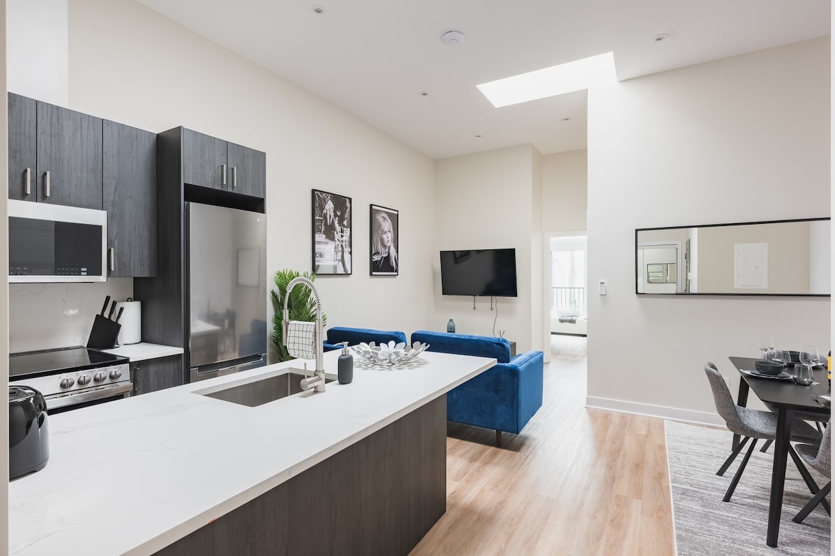 A modern open-concept kitchen features sleek dark cabinetry and a spacious island with a quartz countertop. Natural light pours in through a skylight above, illuminating the stylish dining area with a table set for four, and the living space furnished with a plush blue sofa and a wall-mounted TV.