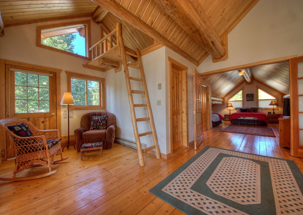 The interior of the cabin features a wooden lofted area accessed by a ladder, with large windows allowing natural light to fill the space. A cozy rocking chair sits near a lamp, and warm wooden flooring enhances the rustic charm.