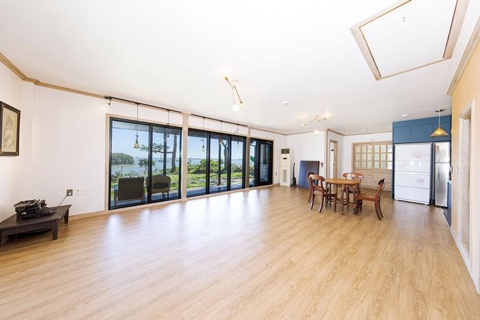 A spacious living area is featured with large windows offering a view of the sea. A wooden dining table with chairs is positioned centrally, surrounded by light-colored walls and wooden flooring. Kitchen appliances are visible in the background, enhancing functionality.