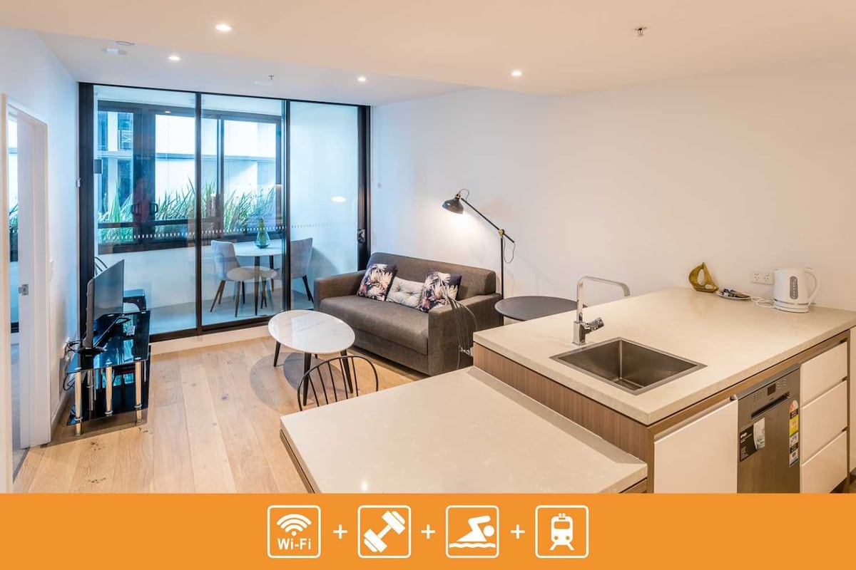 The living room area features a comfortable sofa facing a wall-mounted TV. A small round table and two chairs are positioned nearby, while a modern kitchen with a sink and appliances is visible. Large sliding glass doors lead to a winter-garden balcony filled with greenery.