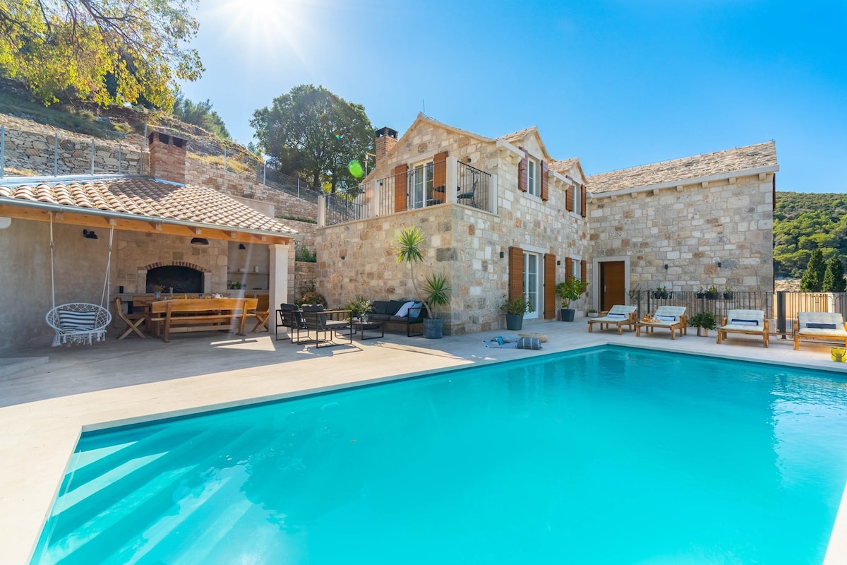 The outdoor area features a private heated swimming pool. A spacious stone villa with wooden window shutters stands alongside a covered outdoor dining area and barbecue, surrounded by lounge seating and sunbeds. Sunlight casts a warm glow over the inviting space.