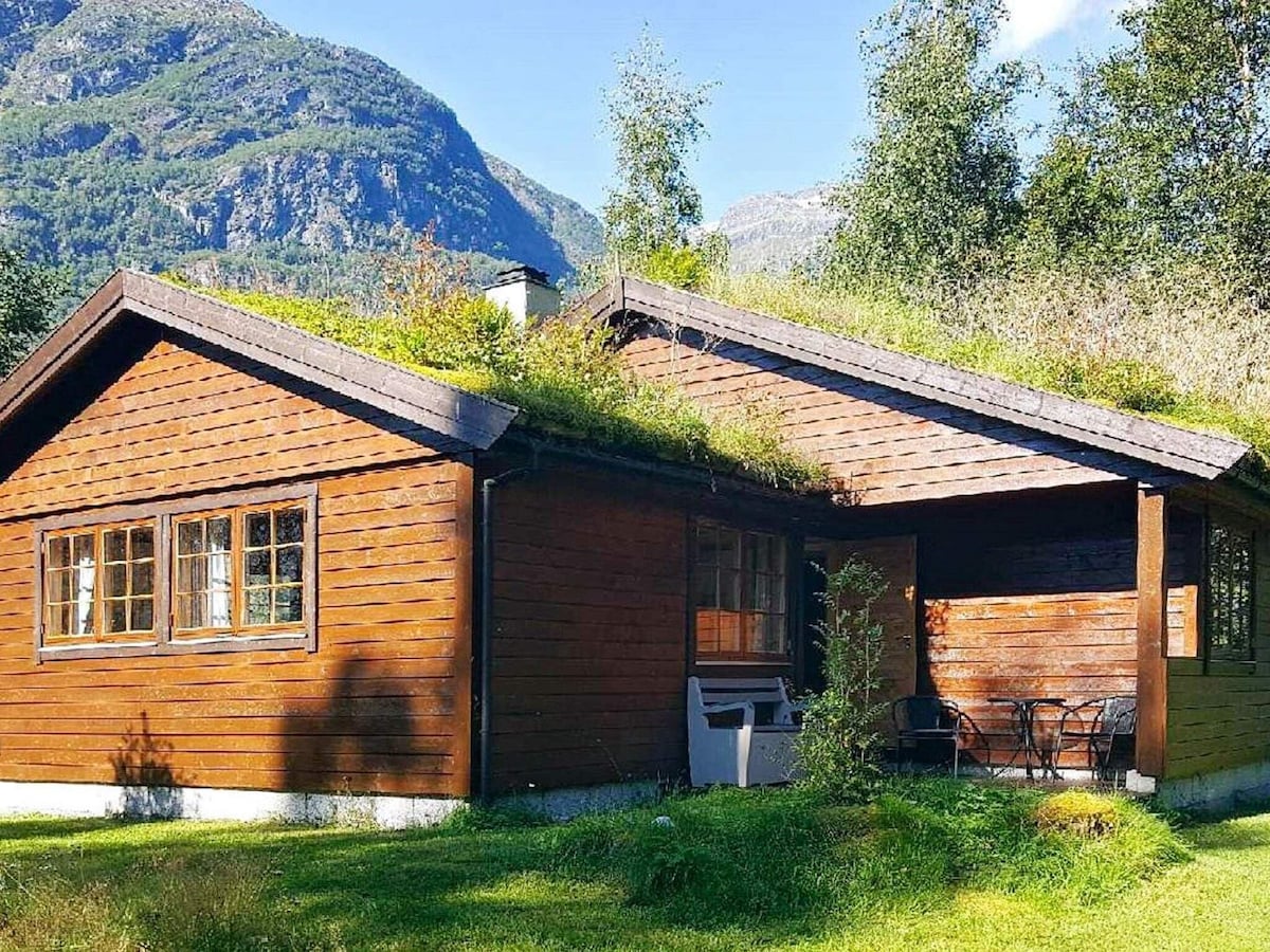 A wooden holiday home with a sloped roof is surrounded by greenery. The exterior features large windows and a covered entrance area. The building is nestled among trees with mountains visible in the background, creating a connection to the natural landscape.