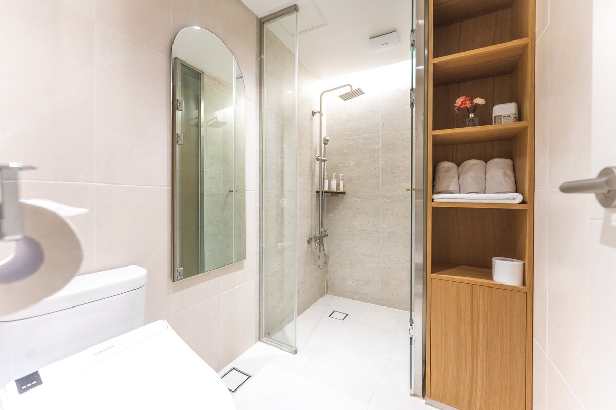 The bathroom features a modern design with a spacious shower enclosed by glass panels. A sleek toilet is positioned beside a wooden shelf displaying neatly stacked towels and toiletries. A full-length mirror reflects the ambient light in the area.