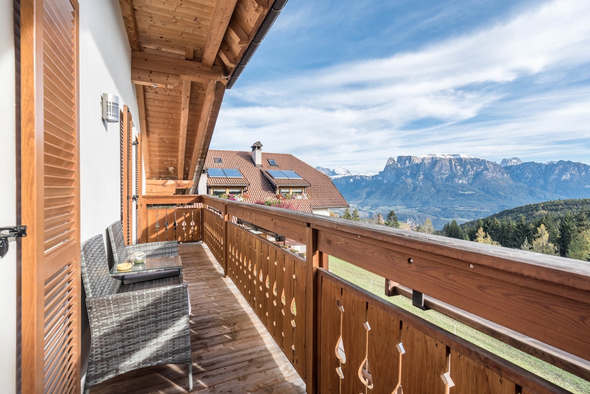 A wooden balcony offers seating with a view of the surrounding mountains. The railing features a decorative design. The distant landscape reveals scenic hills and valleys under a partly cloudy sky, enhancing the outdoor experience.