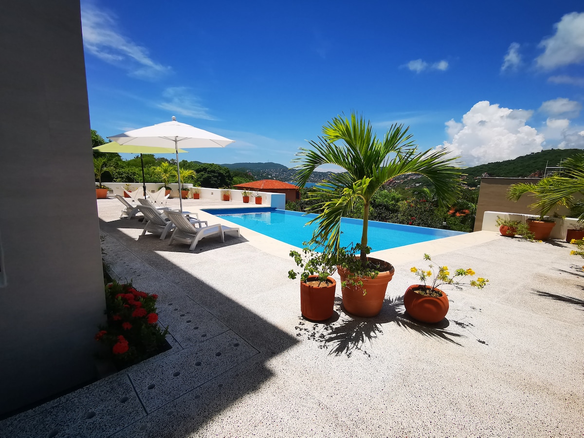An outdoor area features a pristine infinity pool surrounded by lush greenery and potted plants. Poolside loungers and umbrellas provide shaded relaxation. The clear blue sky and distant hills offer a serene backdrop, while vibrant flowers adorn the terrace, creating a tranquil environment.