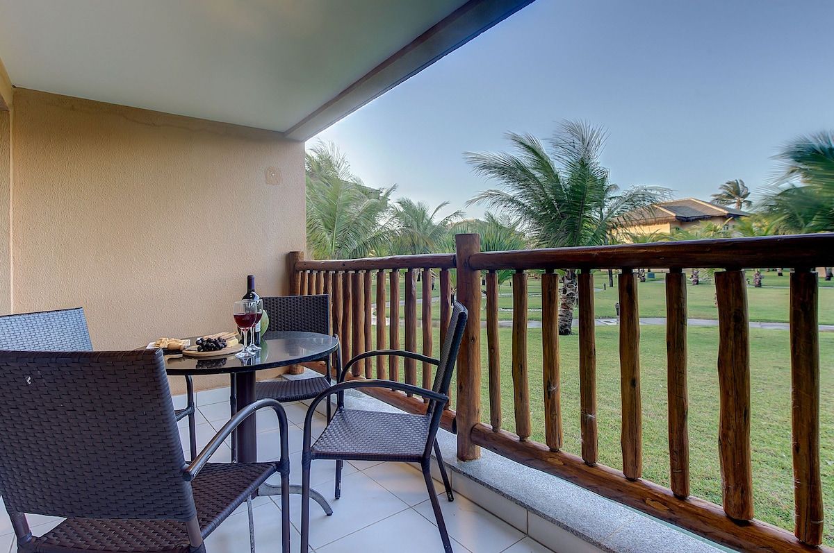 A balcony space is featured, with a round glass table and two woven chairs arranged comfortably. A bottle of wine and two glasses sit atop the table, overlooking lush palm trees and green grass in the distance, creating an inviting outdoor setting.
