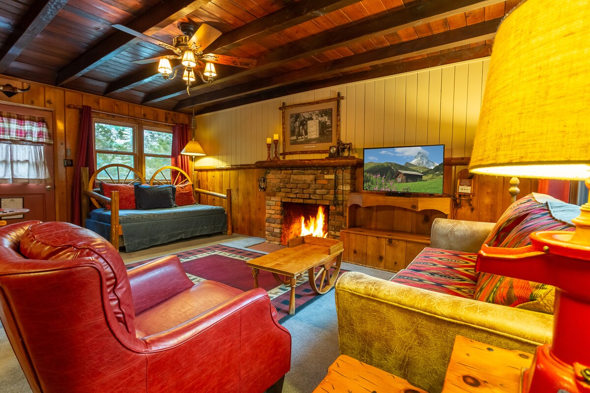 A warm living space is presented with a classic brick fireplace providing cozy ambiance. Comfortable seating includes a red chair and a multi-colored sofa. A wooden coffee table sits at the center, and a television displays a mountain scene, enhancing the rustic decor.