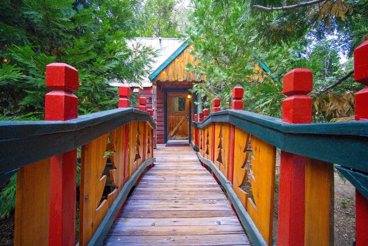 A wooden bridge with red railings leads to a cabin door, surrounded by lush greenery. The cabin features a charming wooden facade with a peaked roof, enhancing the natural setting.