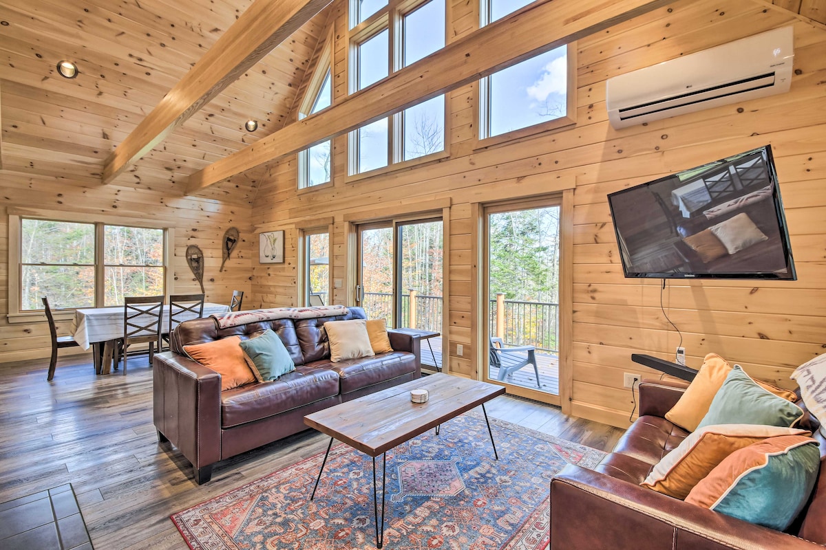 The open living area features vaulted wooden ceilings with large windows, allowing natural light to fill the space. Leather sofas with colorful pillows are arranged around a coffee table, while a Smart TV is mounted on the wall. A dining area is visible in the background.
