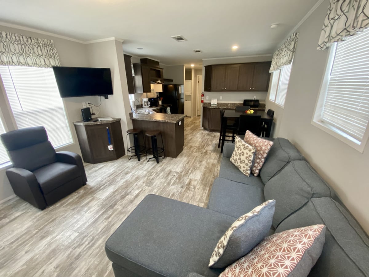 An open living space features a sectional sofa adorned with decorative pillows and a recliner. A kitchen area with dark cabinetry and a breakfast bar creates a functional layout. Natural light comes through the windows, enhancing the bright and welcoming atmosphere.