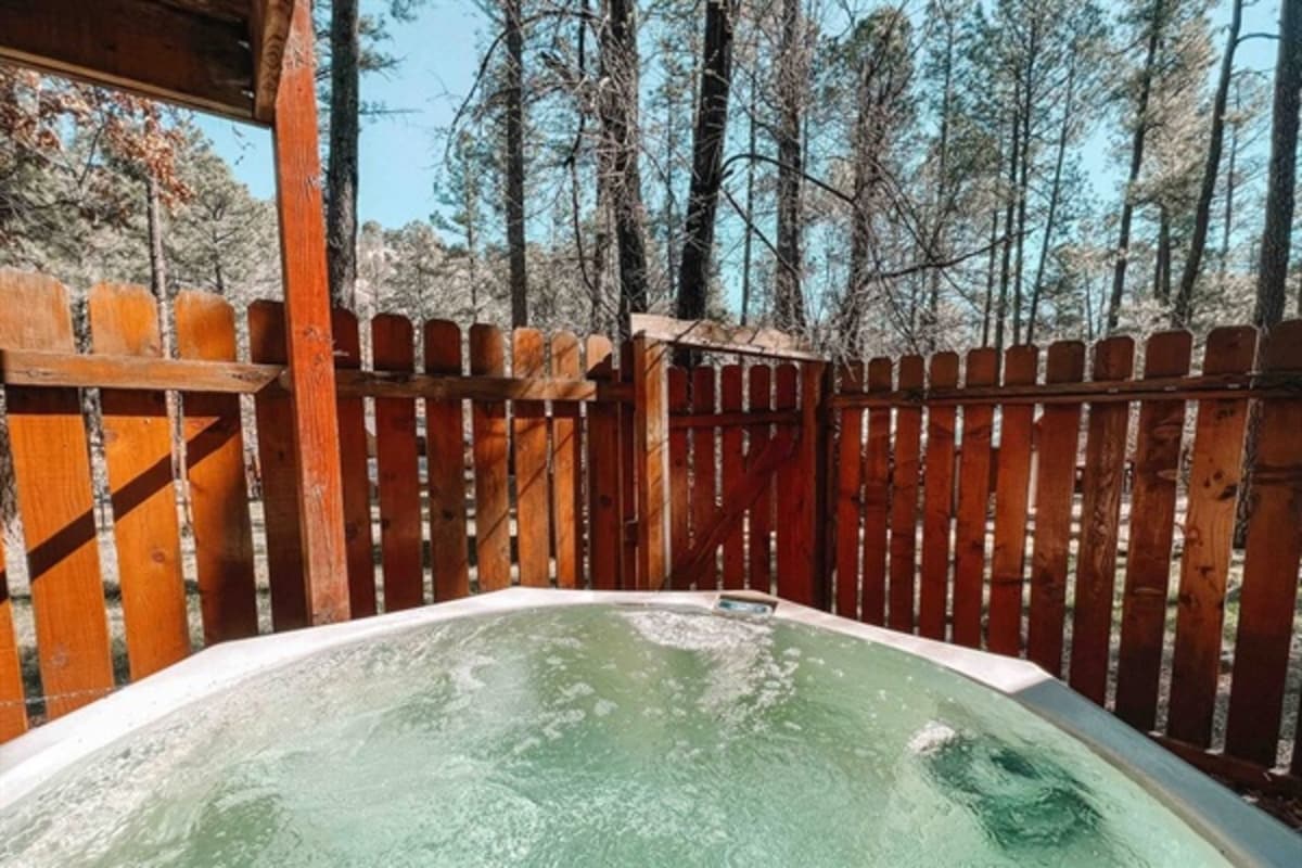 A private hot tub is positioned within a fenced deck area, surrounded by tall trees. The bubbles rise in the water, reflecting the natural light filtering through the forest. Wooden fences provide a sense of seclusion, creating a tranquil outdoor retreat.