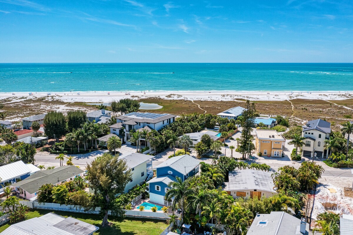 An aerial view captures the vibrant Gulf coastline, showcasing a blend of sandy beaches and lush greenery. Various homes line the streets, with one highlighted by a private pool and ample palm trees, enhancing the coastal ambiance. The clear blue water glistens under the sun.