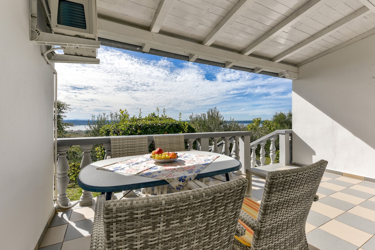 An outdoor terrace is furnished with a round table and four woven chairs, providing a comfortable space. A colorful plate of fruit is placed on the table. The view includes lush greenery and the bay under a partly cloudy sky.