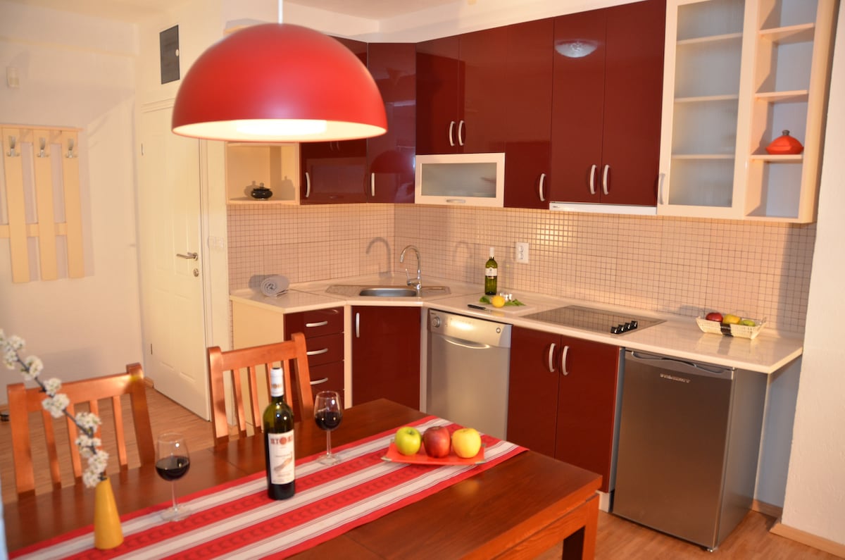 A modern kitchen is equipped with a silver refrigerator and a stainless steel dishwasher. Dark red cabinets enhance the space, complemented by light countertops. A wooden dining table with four chairs is set with glasses of wine, apples, and a decorative table runner.