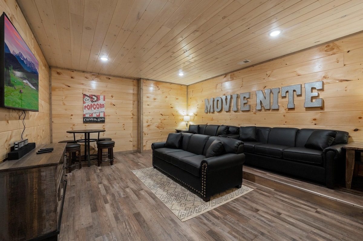 A cozy movie theater room features a dark leather sectional sofa positioned in front of a flat-screen TV. The walls are adorned with wooden paneling, and a decorative sign reads 'MOVIE NITE.' Soft lighting ensures a comfortable atmosphere for gatherings.