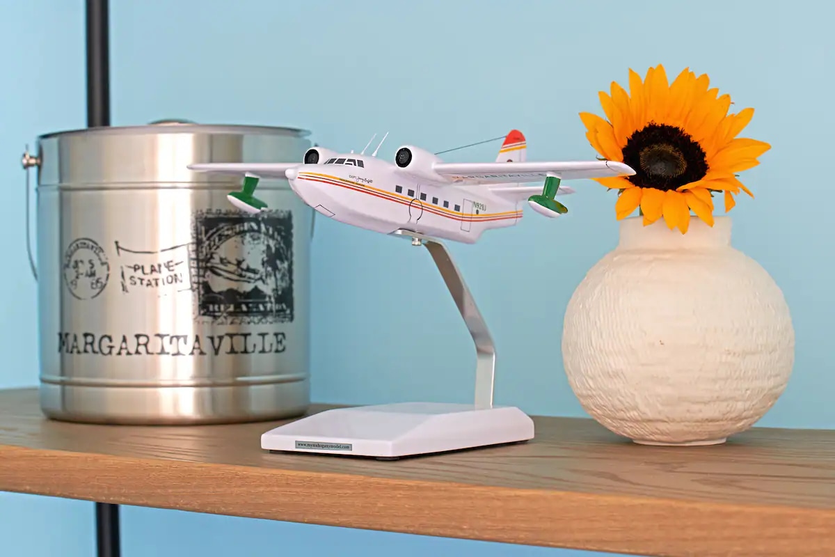 A shelf displays a decorative model airplane on a sleek stand, positioned beside a white, textured vase containing a vibrant sunflower. A metallic container labeled 'Margaritaville' adds a touch of charm, against a soothing blue wall.