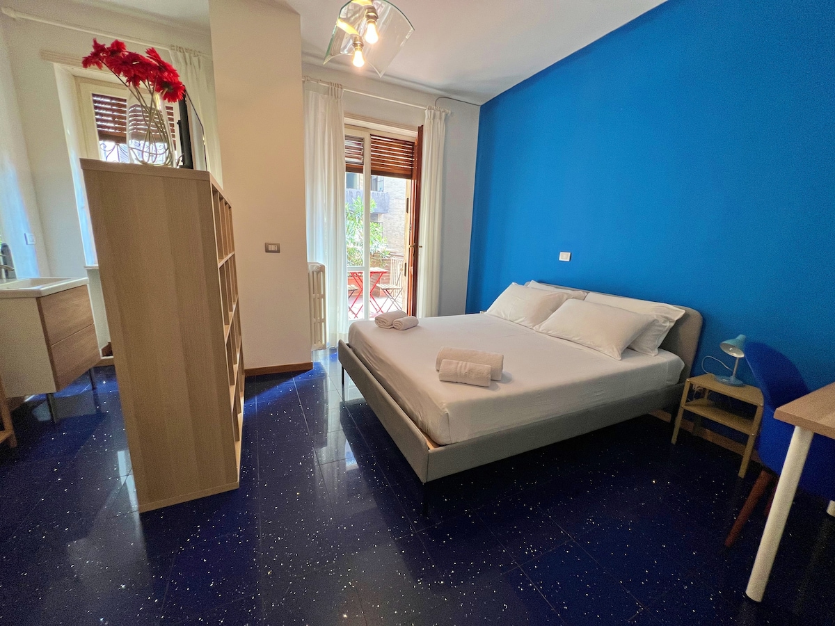 A bright room is enhanced by a vibrant blue wall and natural light flowing through a balcony door. A spacious bed is positioned centrally, while a shelving unit and a desk with a lamp are visible. The glossy floor reflects light, creating a welcoming ambiance.