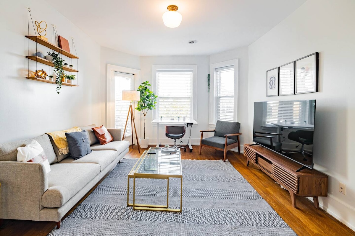 A bright living area features a gray sofa adorned with decorative pillows and a glass coffee table. A corner desk with a chair is positioned near the window, while the TV stands on a wooden media console. Plants and decorative shelves add a touch of greenery to the space.