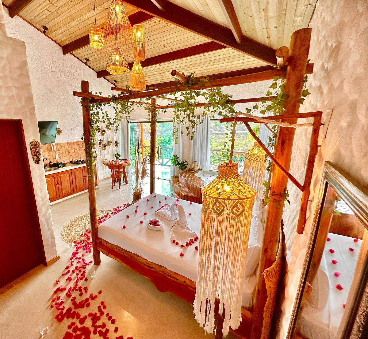 A spacious bedroom features a large bed adorned with floral accents and decorative textiles. Surrounding the bed, natural materials such as wood and rattan provide a rustic touch. Large windows offer a view of greenery, while soft lighting illuminates the space.