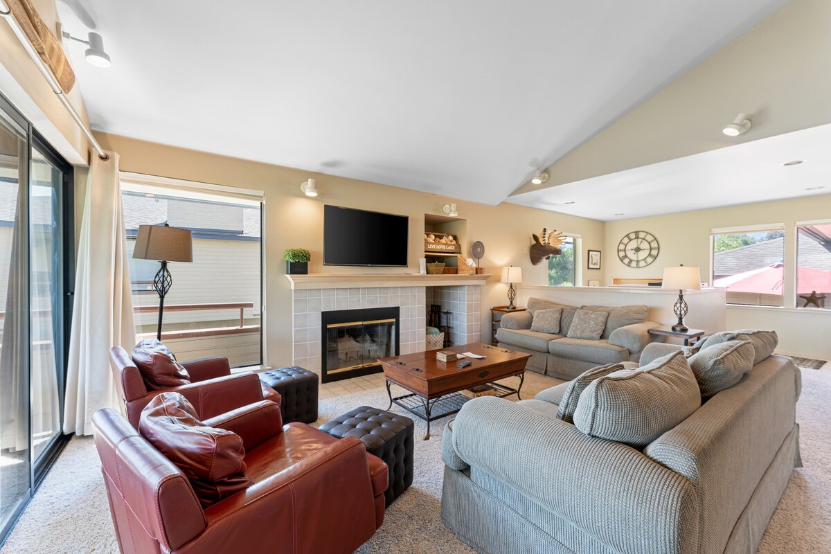 A spacious living area features a cozy layout with multiple seating options, including a large sectional sofa and two armchairs. A stone fireplace is centered beneath a wall-mounted TV, while natural light flows through large windows, creating a bright and inviting atmosphere.