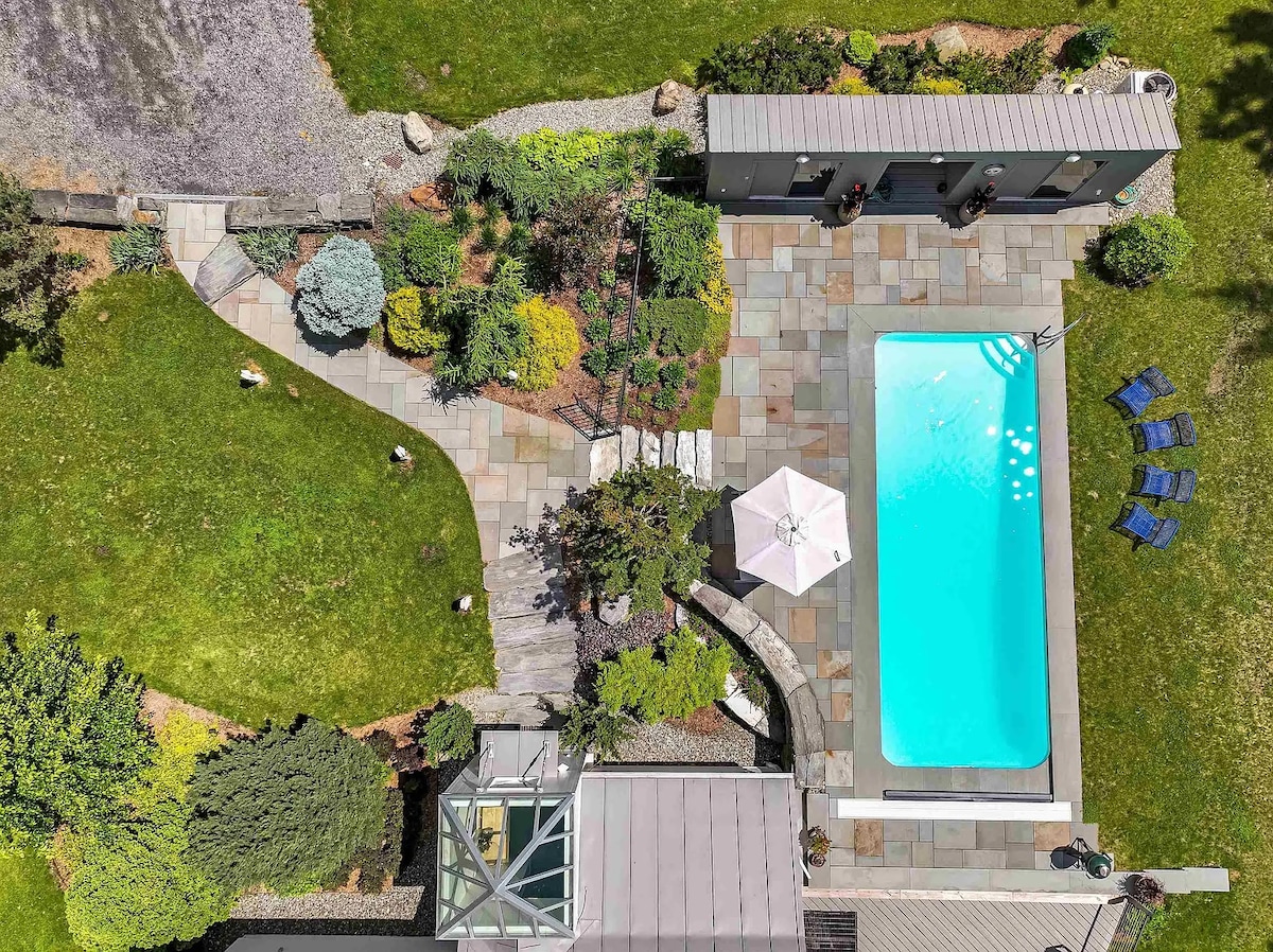 An aerial view of the outdoor area reveals a stone patio surrounding a heated pool, complemented by various landscaping features, including shrubs and trees. A parasol shades a seating area with blue lounge chairs, while a pool house is visible in the background.