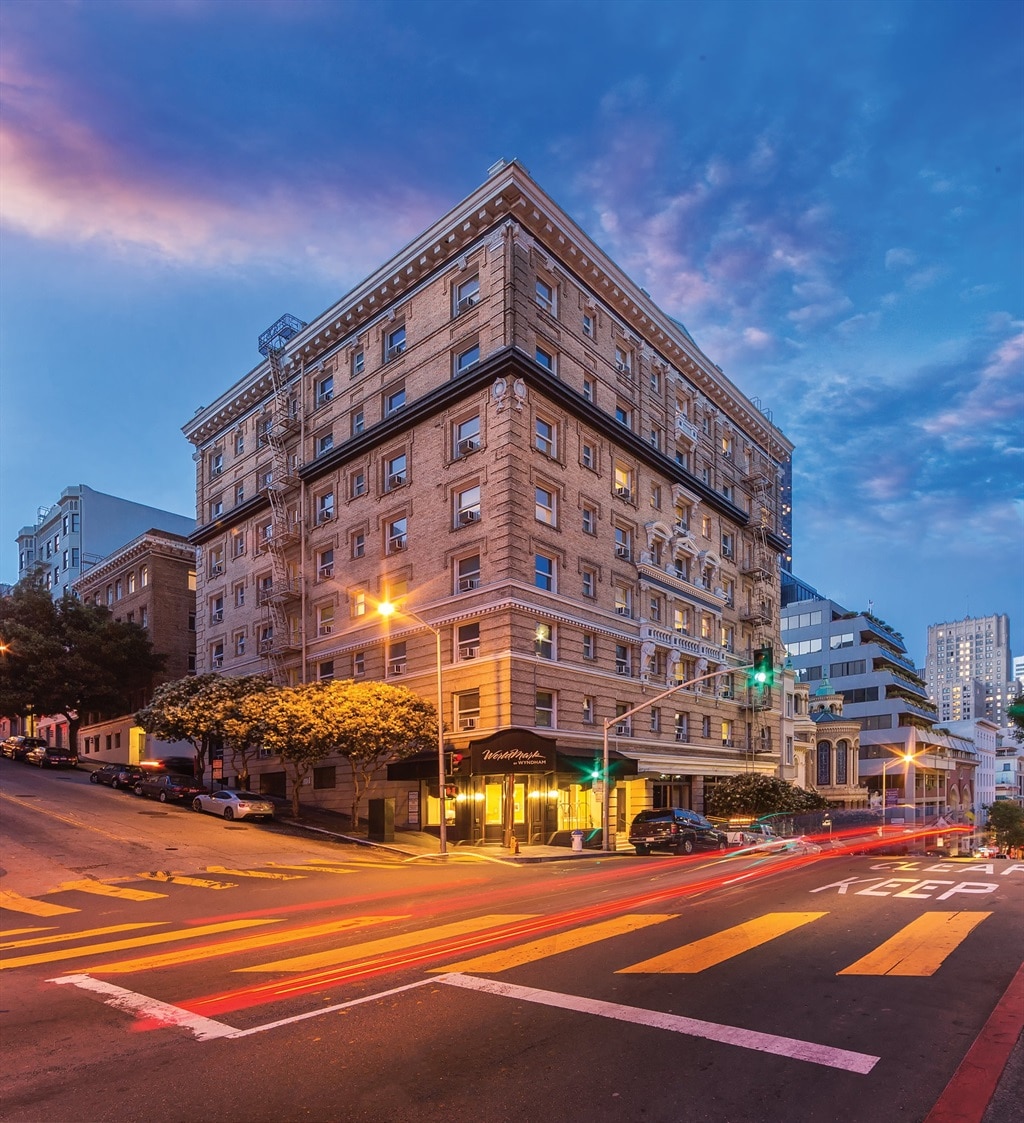 A historic building is depicted from a street view, showcasing a classic architectural style with intricate detailing. Surrounding trees provide a touch of greenery, while evening lights illuminate the street, enhancing the ambiance of the downtown area.