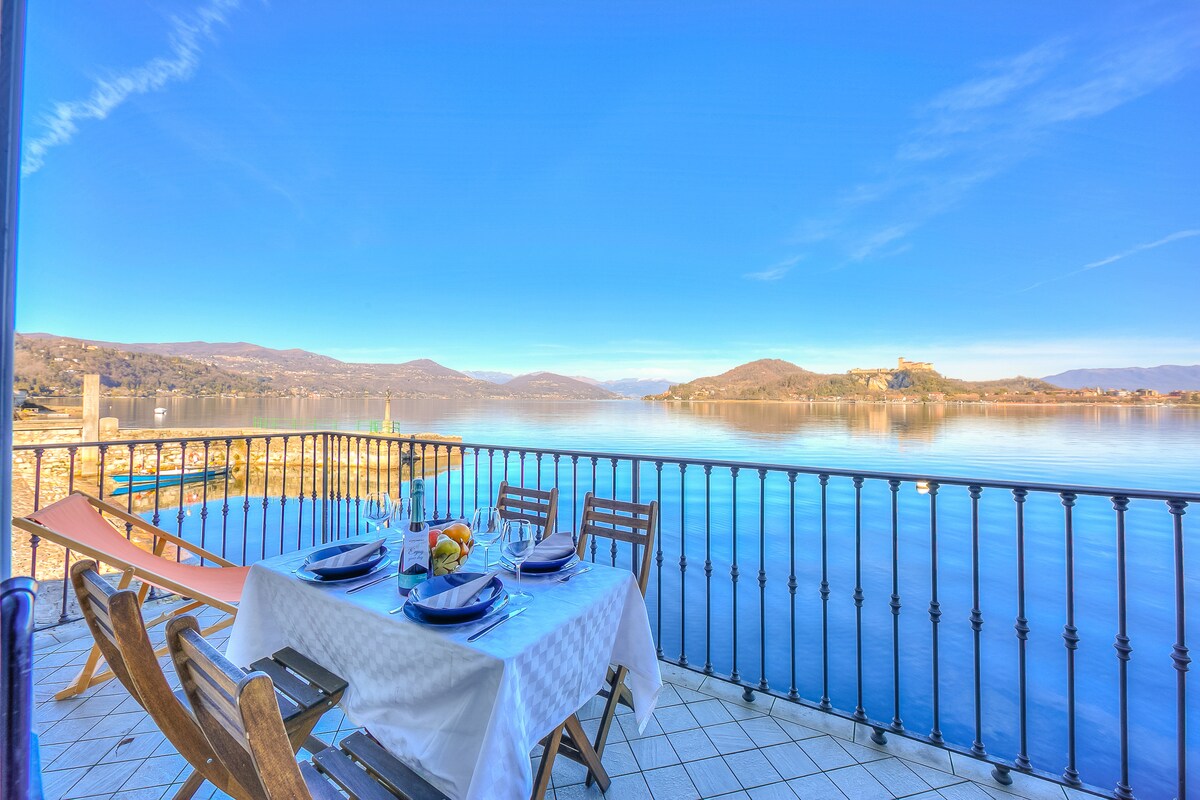 A terrace is shown with a dining table set for a meal, overlooking the tranquil Lake Maggiore. The scene captures stunning views of the lake and hills in the background, framed by a wrought iron railing under a clear blue sky.