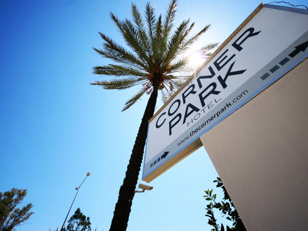 A clear blue sky is visible behind the Corner Park Hotel sign, which features bold black lettering. A tall palm tree stands alongside, providing a tropical feel. Surrounding foliage adds greenery to the scene, creating an inviting entrance to the hotel.