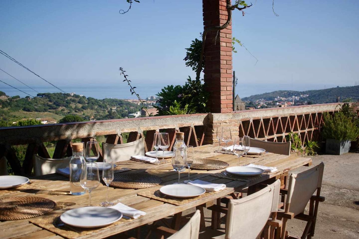 An outdoor dining area is presented with a long wooden table set for a meal, featuring white plates and wine glasses. Surrounding the space are Mediterranean views, including the sea and distant hills, providing a serene backdrop for gatherings.