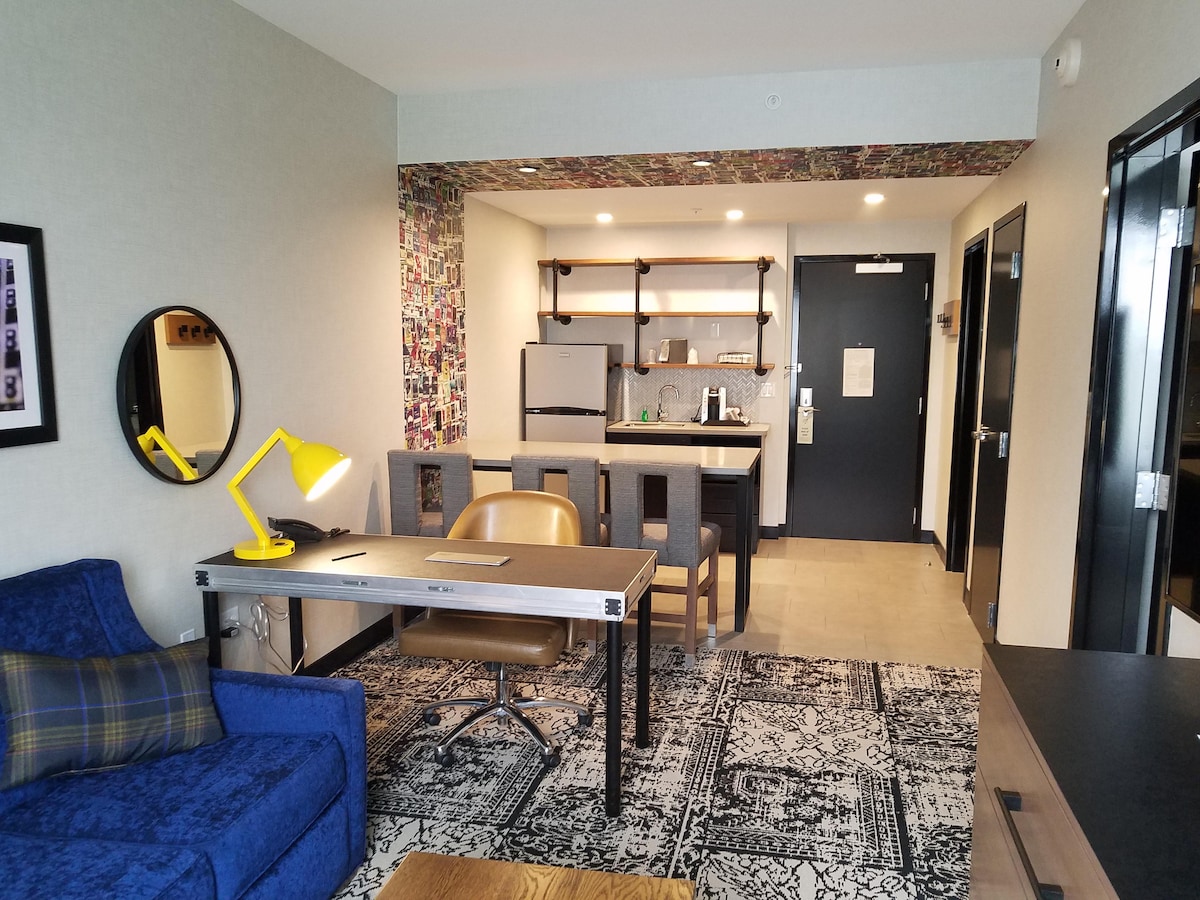 A spacious suite showcases a kitchenette and dining area, accentuated by a colorful feature wall. A large, modern workspace with a golden desk lamp is positioned alongside a comfortable sofa. Natural light enhances the room, creating an open and inviting environment.