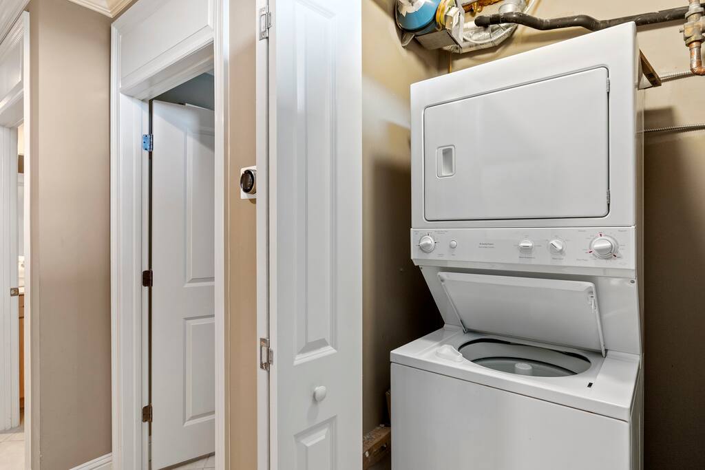 A stacked washer and dryer is positioned in a utility area, featuring a modern white design. The wall is painted in a neutral tone, and a doorway is visible in the background, leading to another room.
