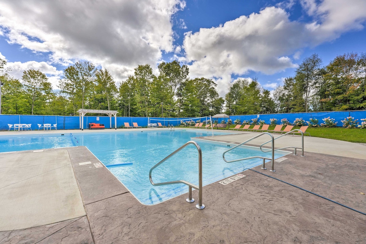 A large outdoor pool is surrounded by a blue privacy wall and lush greenery. Several lounge chairs are positioned around the pool, and clear blue skies are visible overhead, dotted with soft clouds.