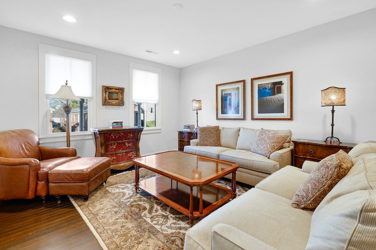 The spacious living area features light-colored sofas and a cozy armchair arranged around a wooden coffee table. Two lamps add soft illumination, while framed artwork decorates the walls. Natural light filters through two windows, enhancing the inviting atmosphere.