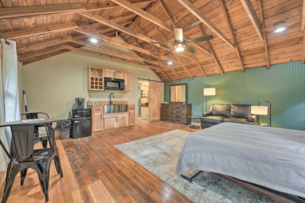 A studio cabin interior is presented with vaulted wooden ceilings. The space includes a cozy living area with a leather sofa, a kitchenette with wooden cabinetry, and a queen Murphy bed on a polished wooden floor. A small dining table and black chairs are viewed near a window.
