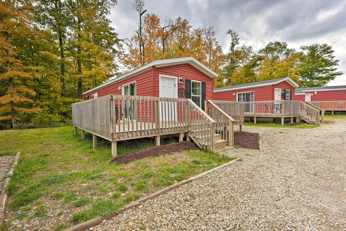 A charming cabin with a red exterior is positioned amidst trees, featuring a spacious wooden deck complemented by railings. A gravel pathway leads to the entrance, inviting guests to explore the natural surroundings.