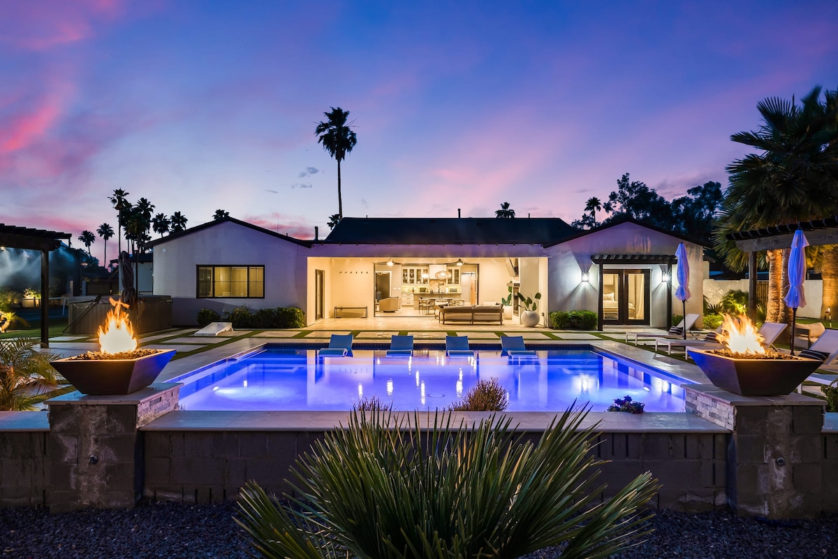 The inviting exterior of the property features a spacious pool surrounded by lounge chairs and accentuated by glowing fire bowls. The home is elegantly backlit by the sunset, highlighting the seamless indoor-outdoor living space with large glass doors that connect the patio and kitchen.