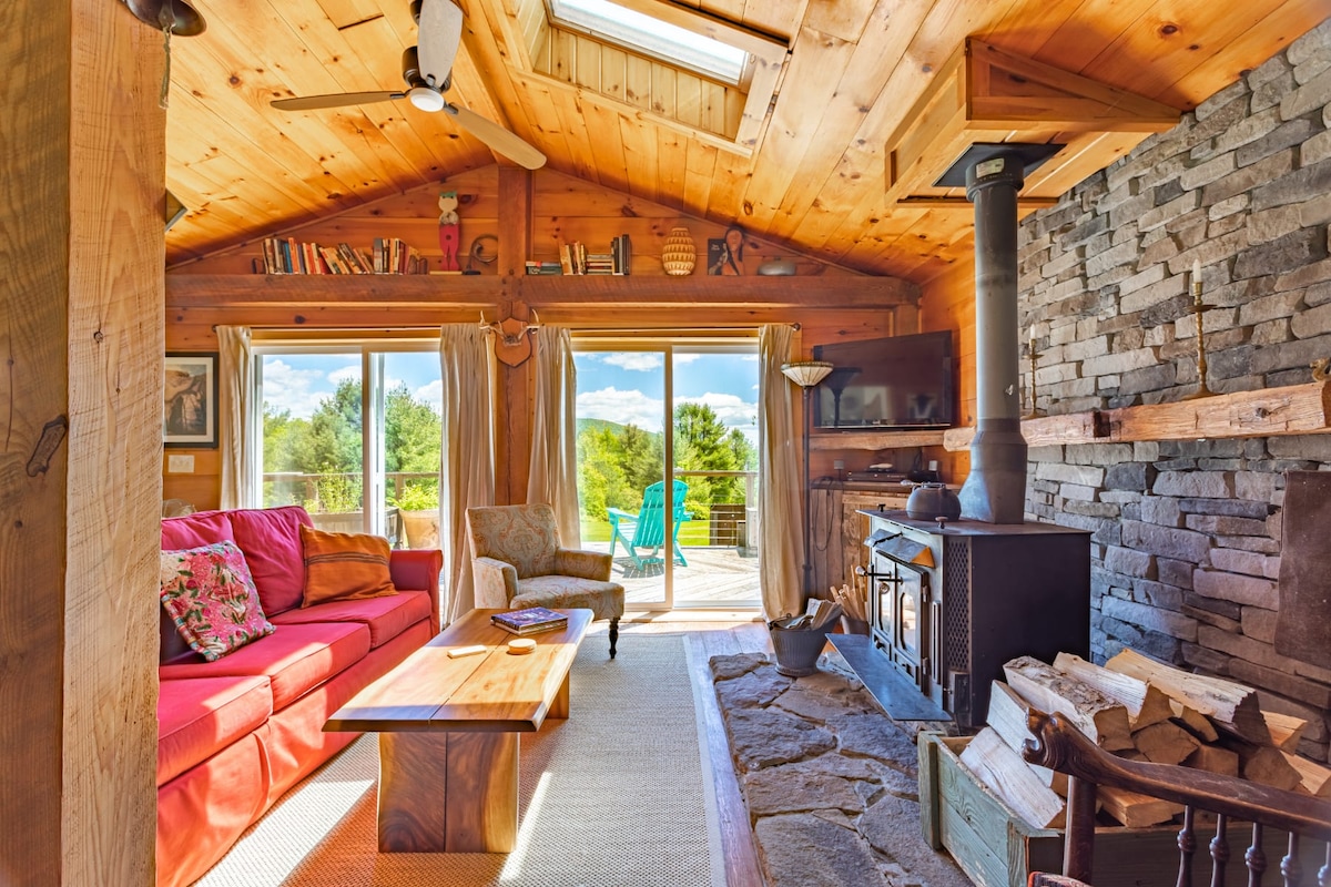 A warm living area features wood paneling and a cathedral ceiling, with large glass doors providing access to the deck. A red sofa and a patterned chair offer comfortable seating, complemented by a rustic coffee table. A stone fireplace adds a cozy focal point to the room.